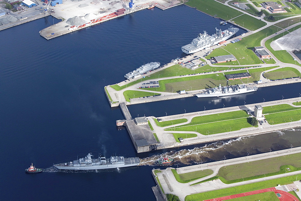 Aerial of tug boat towing battleship out of loch, naval harbour at Wilhelmshaven, Lower Saxony, Germany