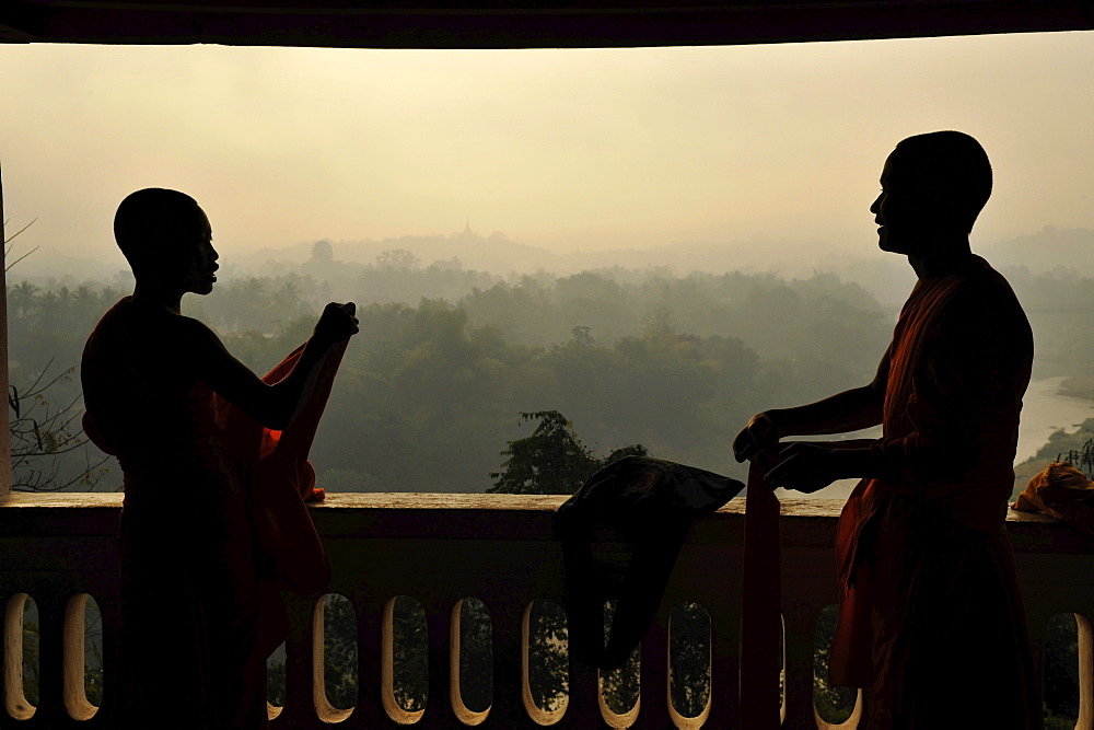 Buddhist monks, novices, cladding their robes, view to the East over trees and hills in morning dust, Phu Si hill, Luang Prabang, Laos