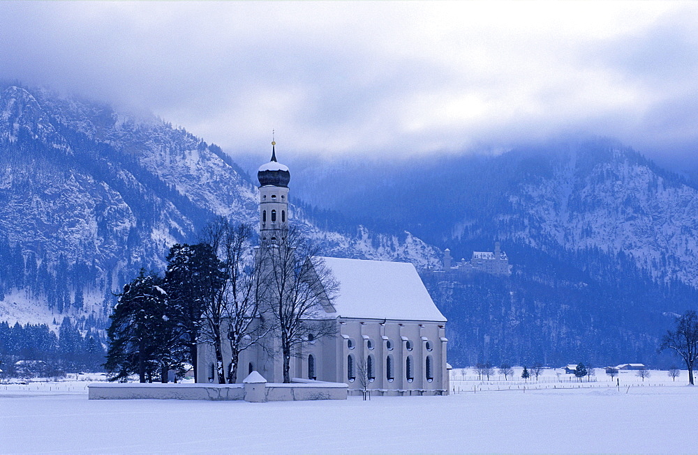 Europe, Germany, Bavaria, Schwangau near Fuessen, St. Coloman pilgrimage church surrounded by trees and Neuschwanstein Castle in the mountains