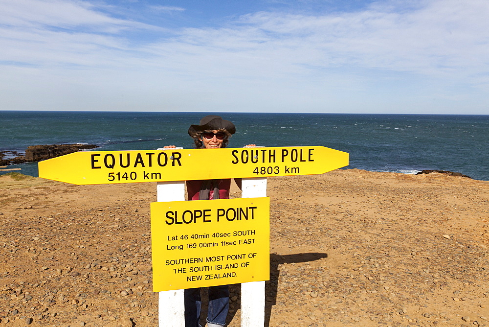 Woman behind a sign showing the southernmost point of South Island, Slope Point, Catlins, South Island, New Zealand