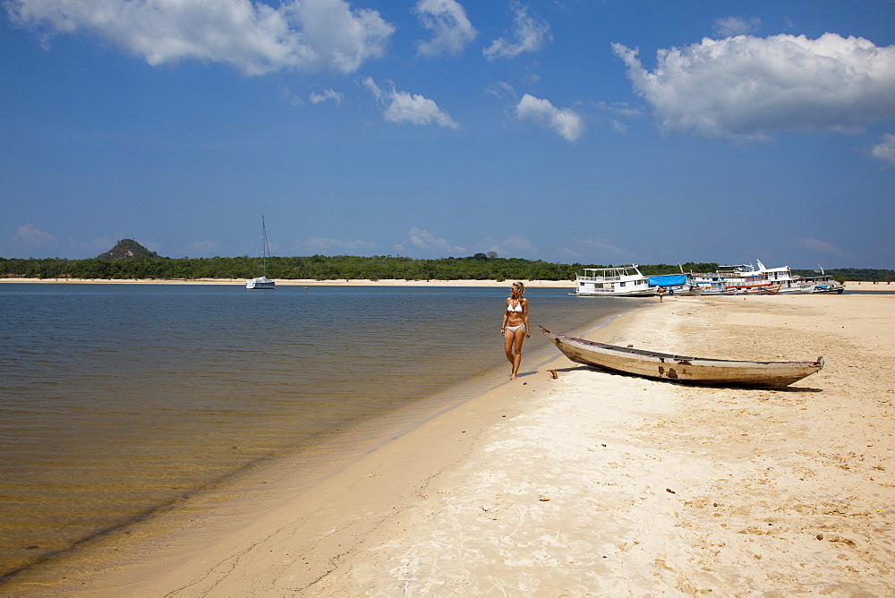 Woman strolling along the beach on a side arm of the Amazon river with Amazon river boats in the distance, Alter do Chao, Para, Brazil