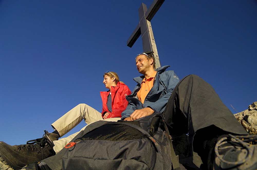 Couple on a hiking tour at heimgarten, having a rest at the summit cross, Upper Bavaria, Bavaria, Germany