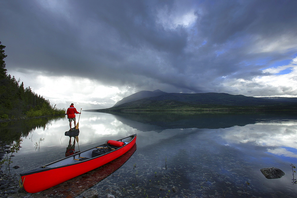 Man Standing on Rock under Storm Clouds beside a Canoe, Kusawa Lake, Yukon