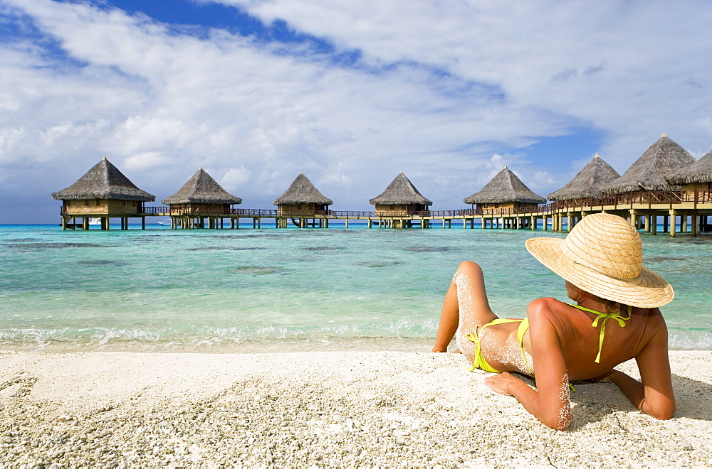 French Polynesia, Tuamotu Islands, Rangiroa Atoll, Woman lounging on beach, Luxury resort bungalows in background.