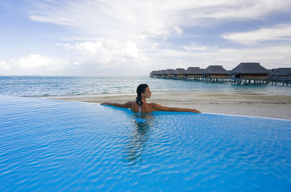 French Polynesia, Moorea, Woman relaxing in resort pool, Luxury resort bungalows in background.