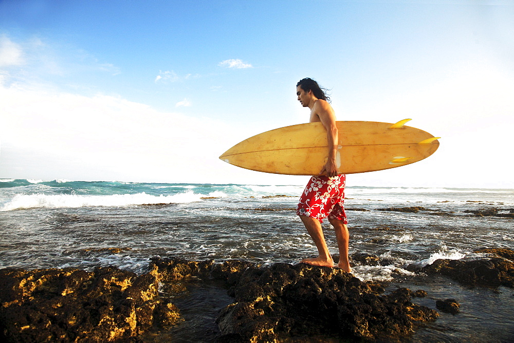 Hawaii, Oahu, young man walking on rocky coastline with surfboard