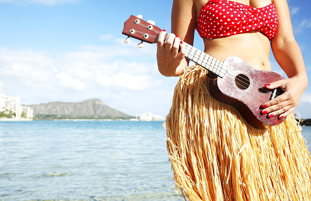 Hawaii, Oahu, Waikiki, Hula dancer playing ukulele along beach - close-up of midriff, Diamond Head in the distance.