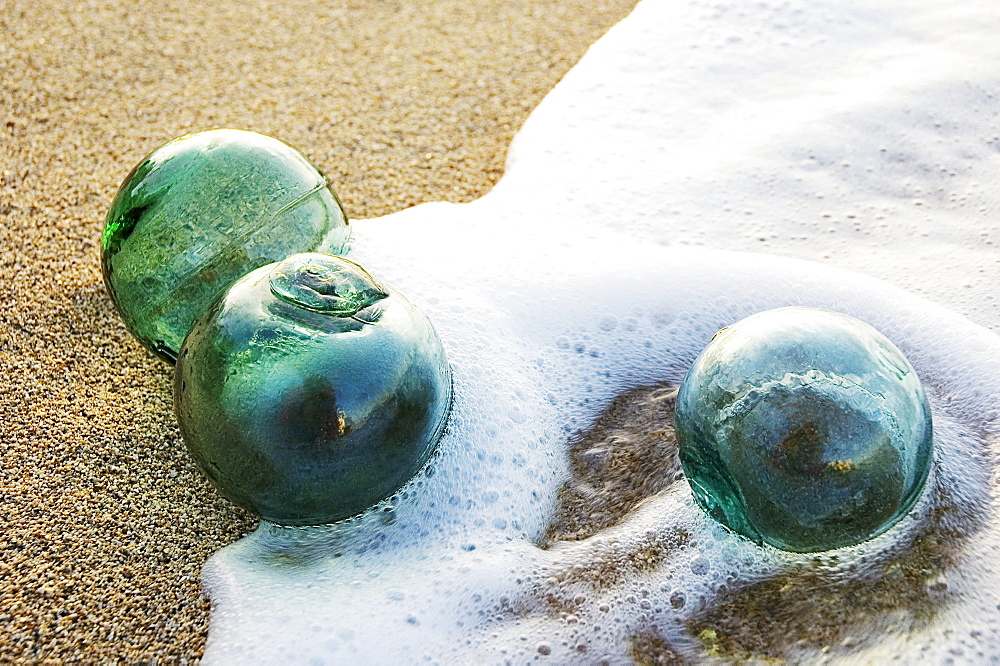 Three glass fishing floats roll on the sandy shoreline with ripples of water and seafoam