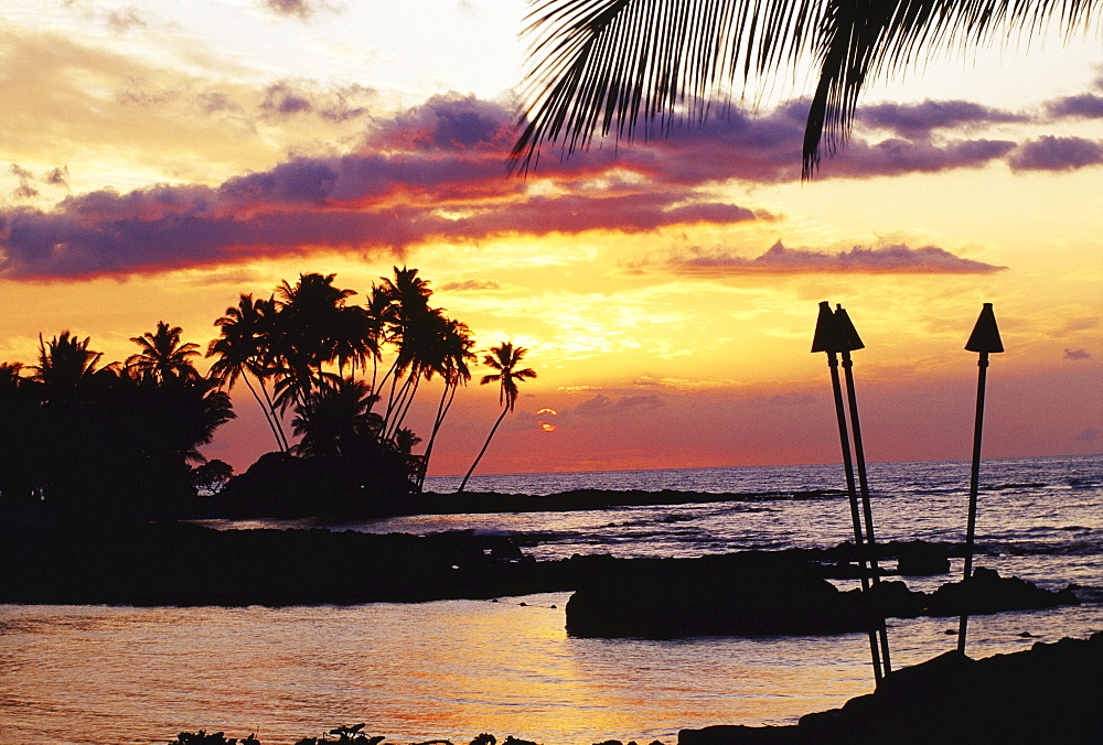 Hawaii, Big Island, Kohala, Waiulua Bay, Orange sunset with palm trees and tiki torches.