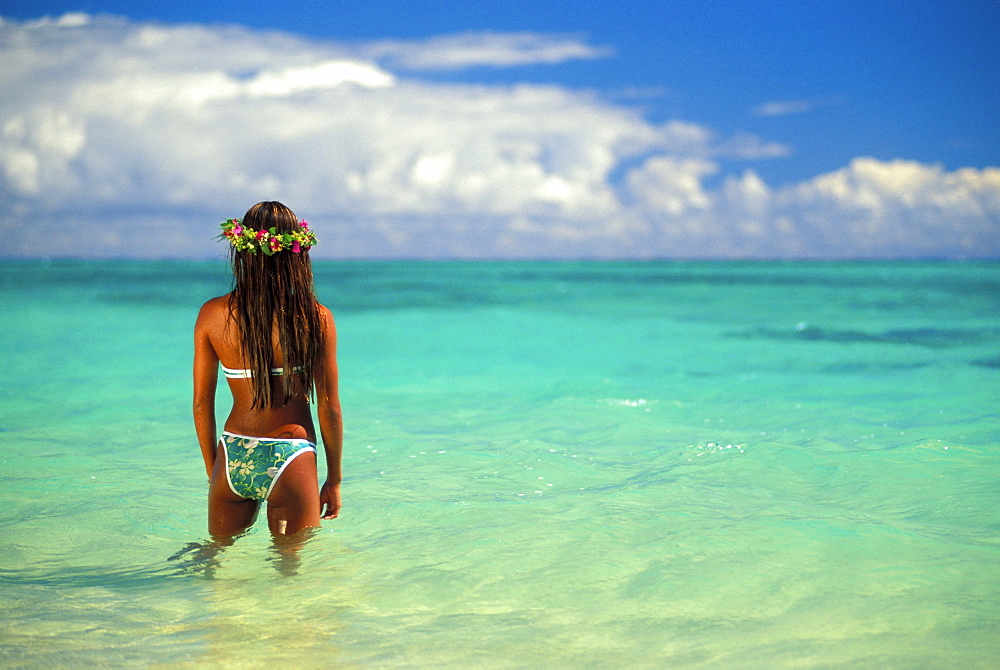 Back view of woman in bikini, stands in shallow turquoise water, haku lei