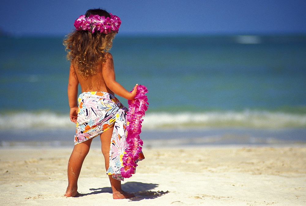 Back view of little girl at the beach, holding pink lei and wearing a pareo