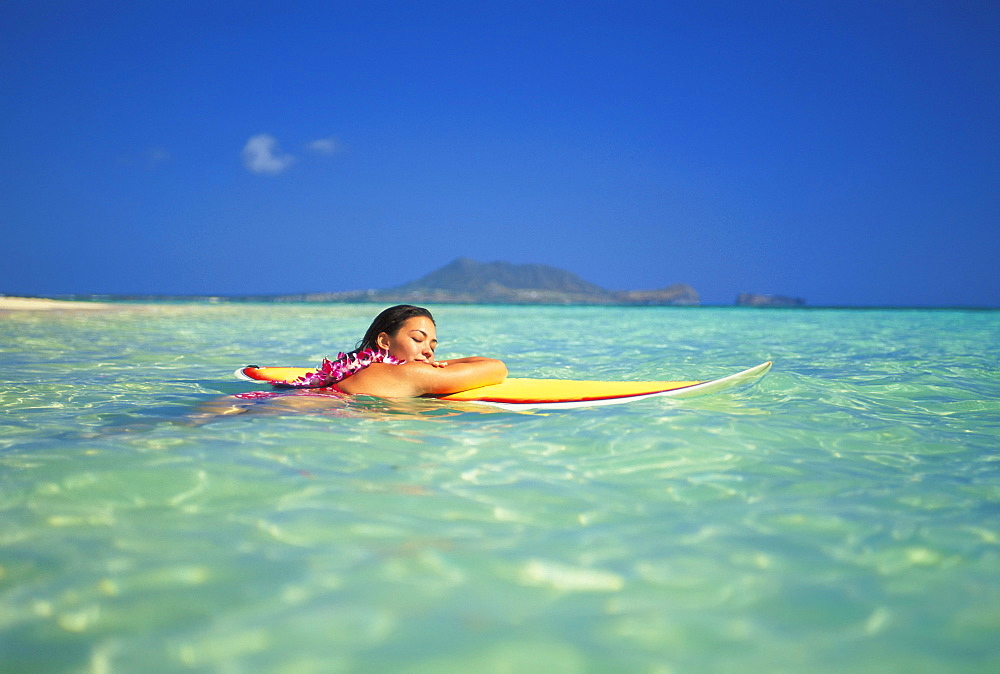 Hawaii, Oahu, Lanikai, Woman eyes closed resting head/arms on surfboard on clear water.