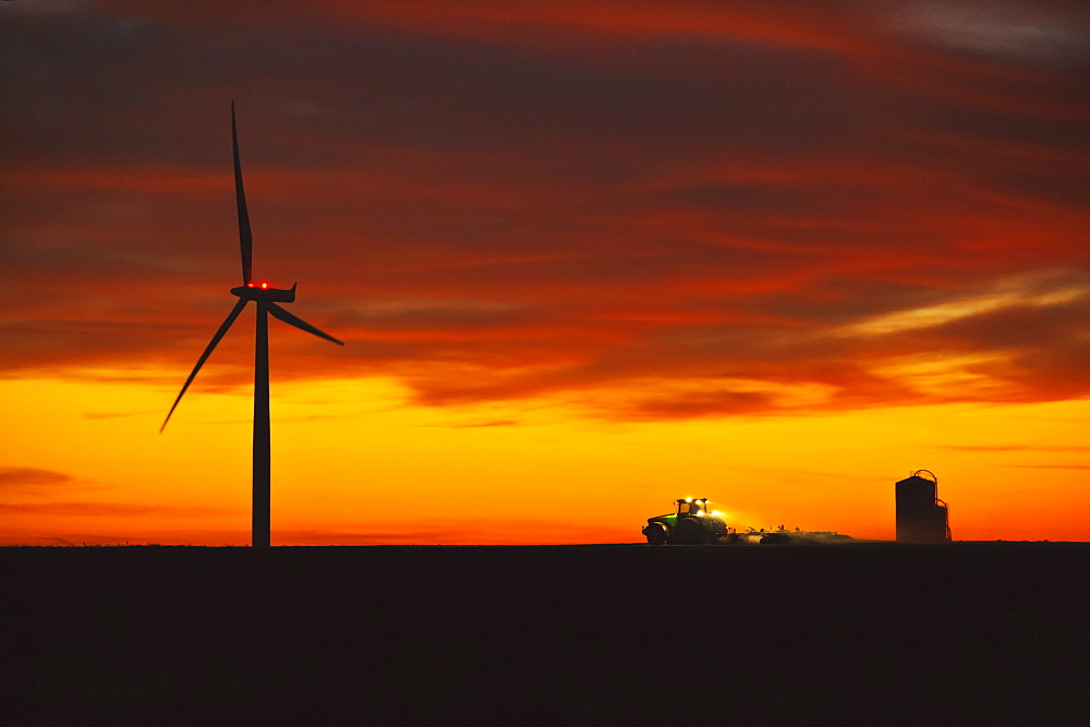 Agriculture - A farmer tills his field at dusk after corn harvest in late Autumn, below an electric wind turbine, illustrating the coexistence of traditional and modern or