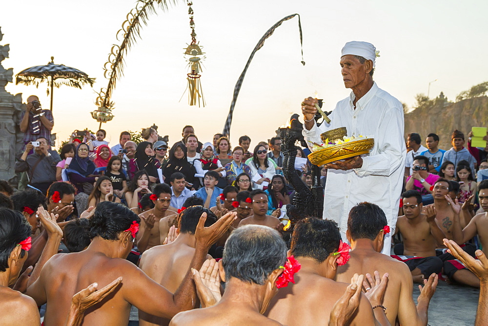 Priest carrying incense and water for blessings surrounded by men sitting in a circle chanting in trance during a Kecak dance performance, Ulu Watu, Bali, Indonesia