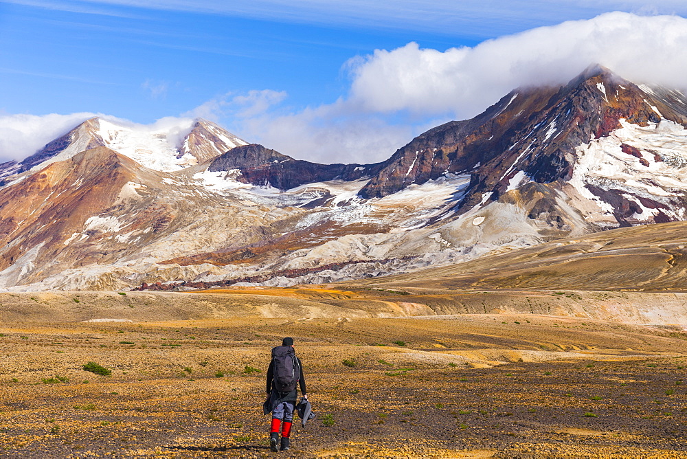 A backpacker crosses the exotic landscape of the ash and pumice-covered Valley of Ten Thousand Smokes in Katmai National Park, with Mt. Katmai (left), Trident Volcano (right), and the Knife Creek Glaciers looming in the distance; Alaska, United States of