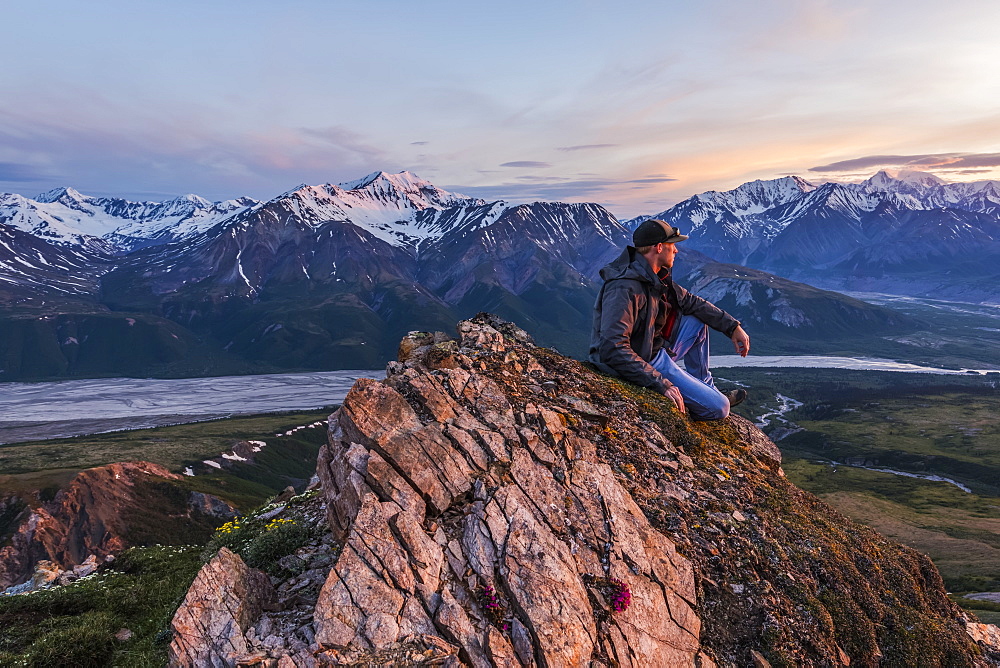 A man observes a tranquil sunset from an alpine perch in the Alaska Range high above the Delta River; Alaska, United States of America