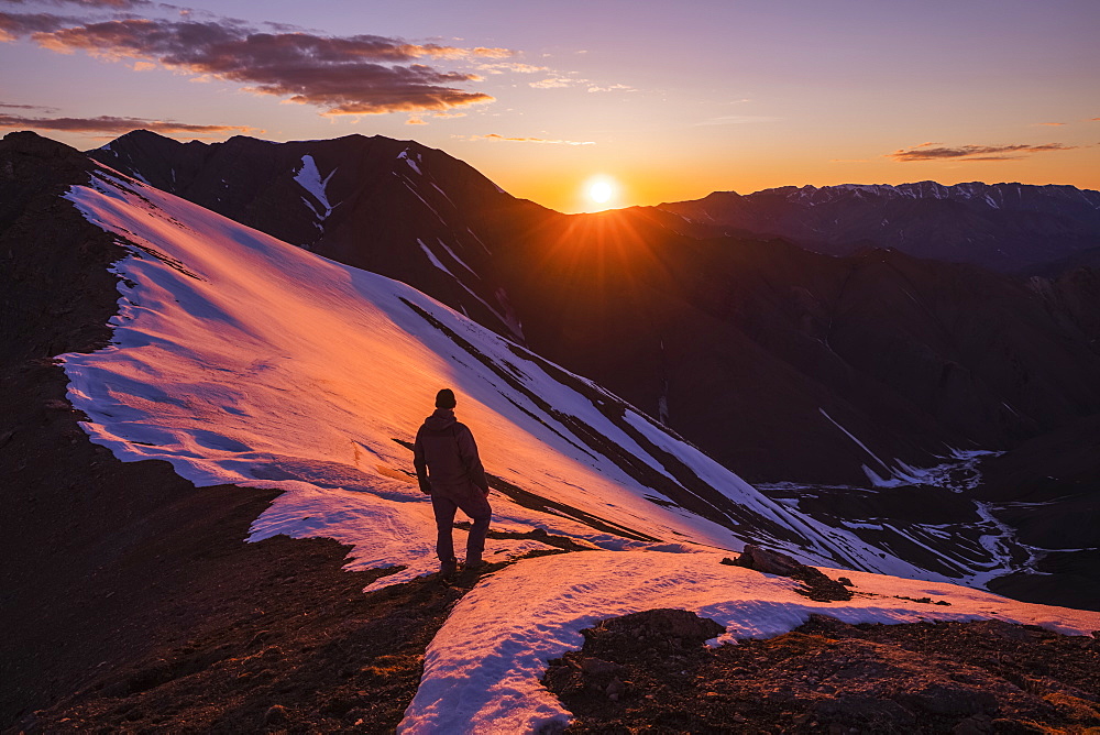 A man stands silhouetted on a ridge as the sun sets behind Polychrome Mountain in Denali National Park in early summer; Alaska, United States of America