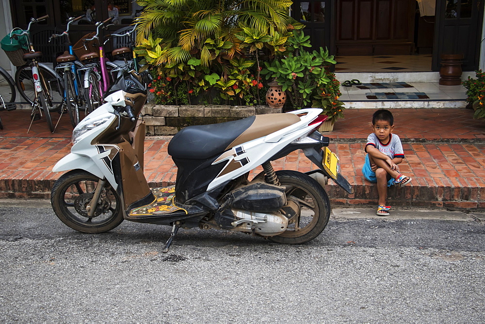 Boy sitting along Sisavangvong Road next to a parked motorcycle, Luang Prabang, Luang Prabang, Laos