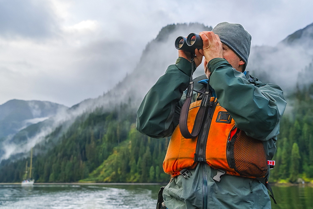 Man looking through a pair of binoculars with his sailboat in the distance, Great Bear Rainforest, Hartley Bay, British Columbia, Canada