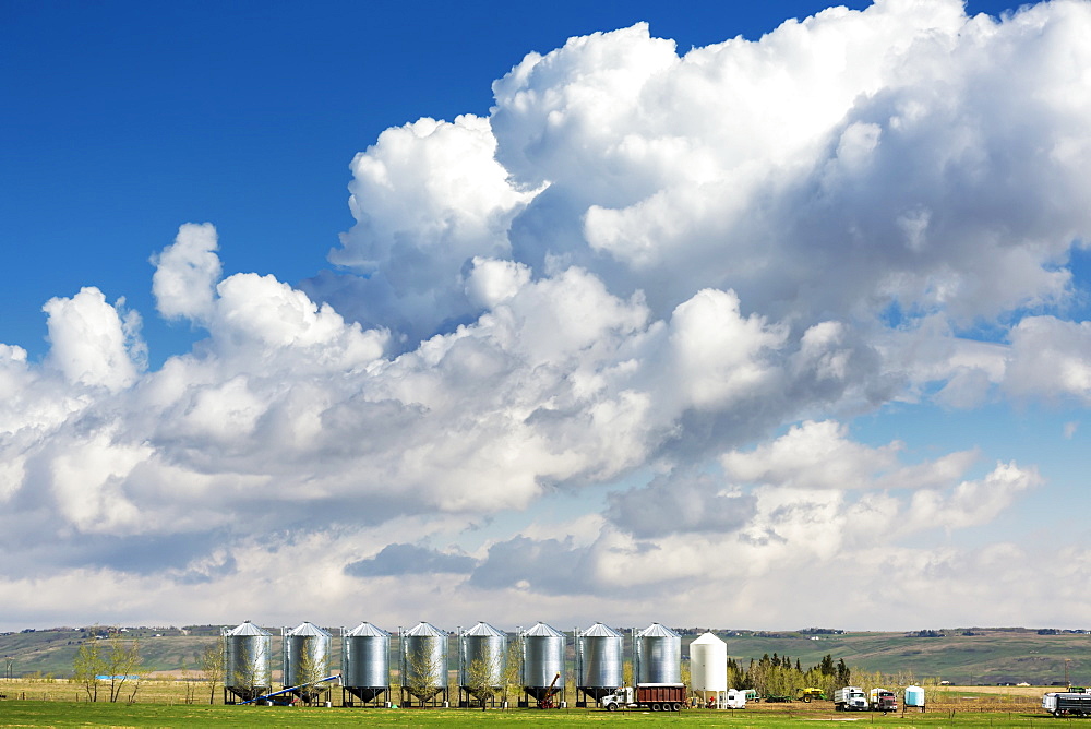 A row of large metal grain bins with dramatic storm clouds and blue sky in the background, West of Calgary, Alberta, Canada