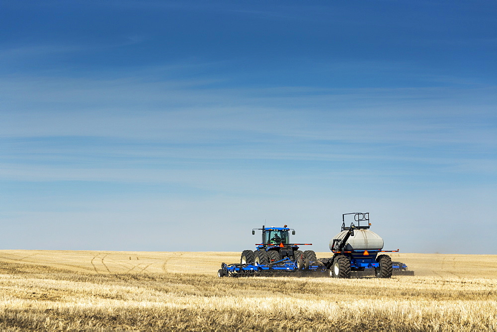 Tractor with air seeder in field with blue sky and hazy clouds, near Beiseker, Alberta, Canada