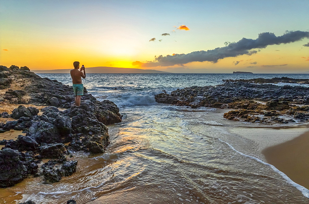 A young man in a bathing suit stands on the volcanic rock on the shore capturing the golden sunset with a smart phone camera, Kihei, Maui, Hawaii, United States of America