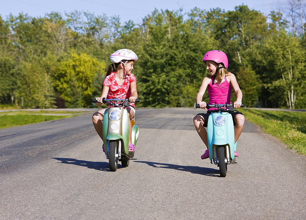 Two girls riding electric scooters down a paved country road, Canada