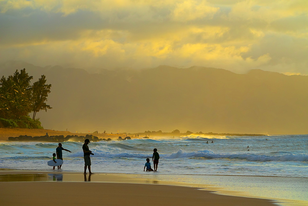 Silhouette of surfers on Kelki Beach with dramatic glowing clouds above at sunset, Oahu, Hawaii, United States of America