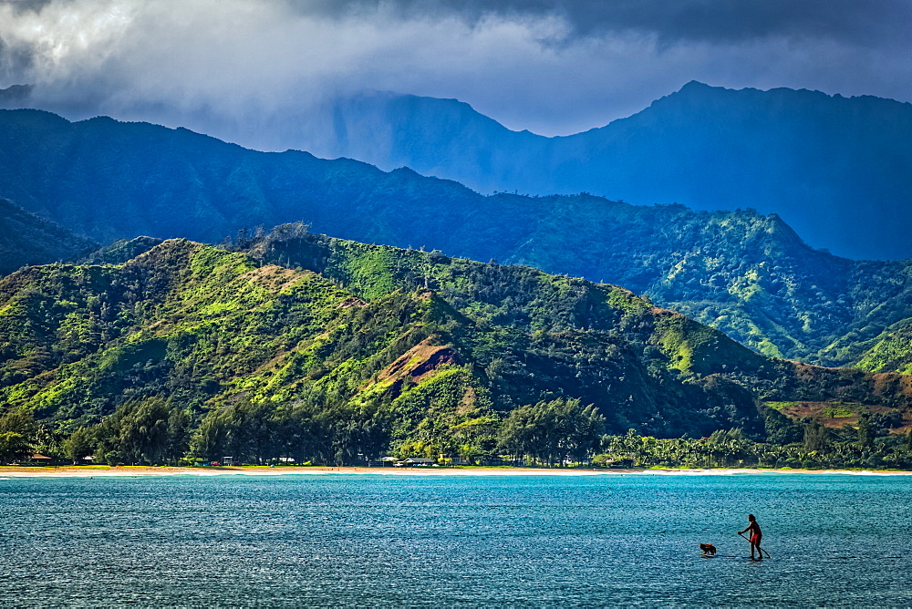 A man and his dog on a stand up paddle board in Hanalei Bay with mountains in background, Hanalei, Kauai, Hawaii, United States of America