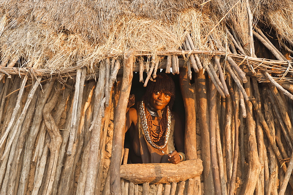 Pregnant Hamar woman with necklaces made of cowry shells coming out of her wooden hut, Omo River Valley, Southern Ethiopia, Africa