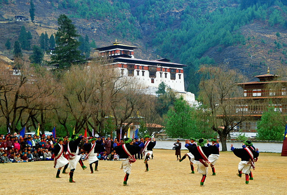 A crowd sit to watch men dressed in traditional costumes dancing as part of a dance festival taking place beside the Paro Dzong a fortress and buddhist monastery, Paro, Bhutan.