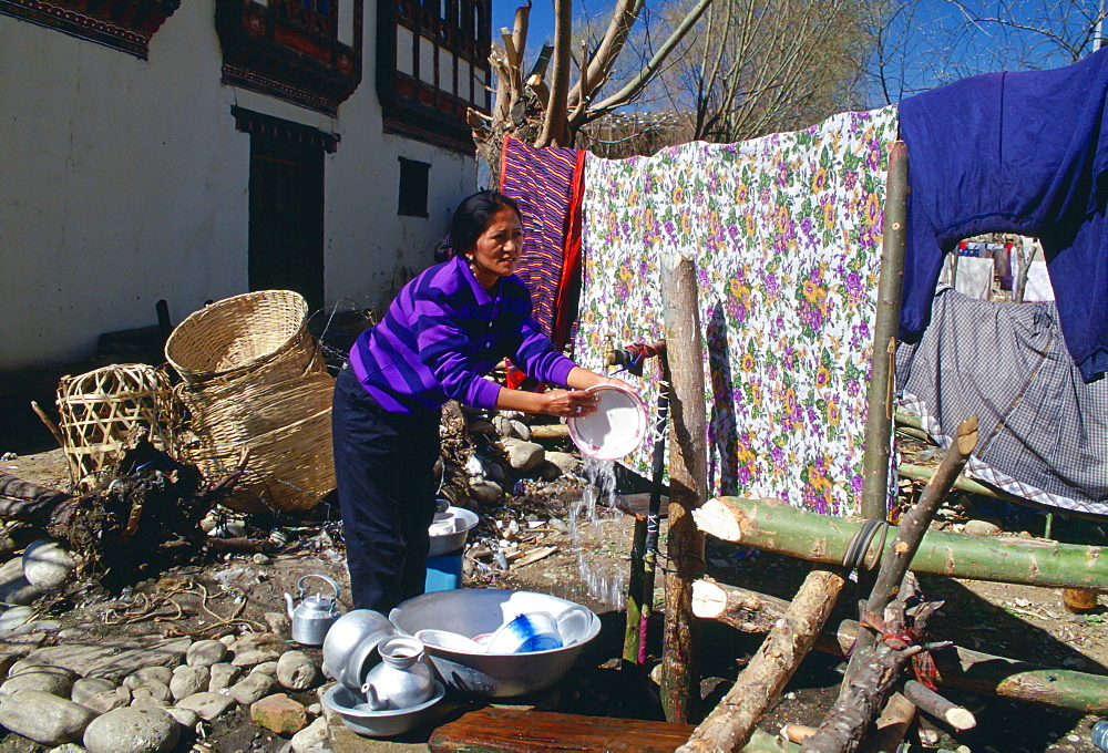 Domestic chores without modern electrical appliances - a woman washing up by hand using cold water from an outdoor tap.  Street scene in Bhutan.