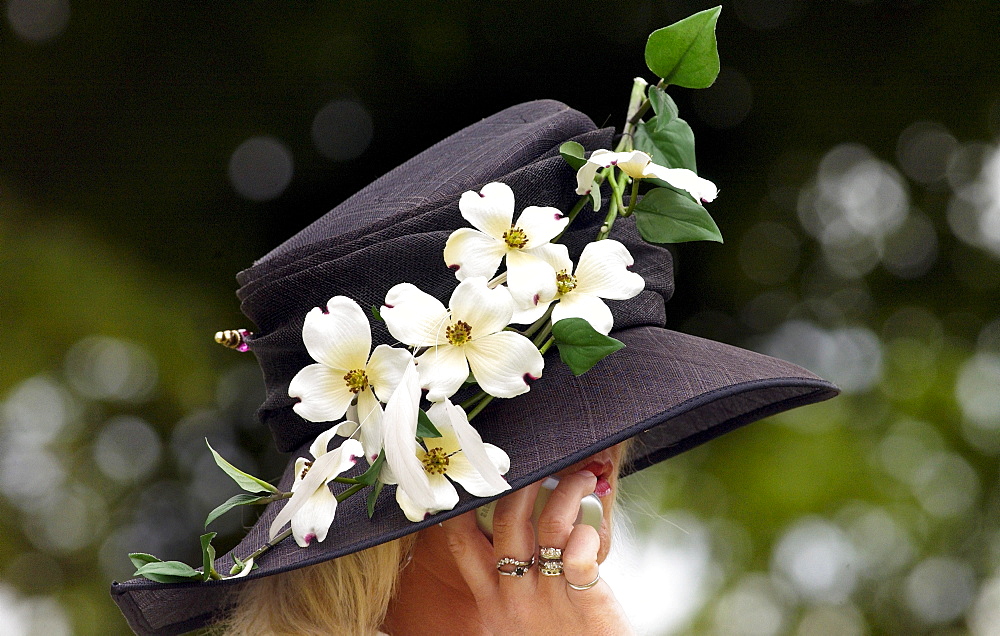 Race-goer wearing a hat with flowers in typical Ascot fashion at Royal Ascot Races. She is taking a call on her mobile cell phone.