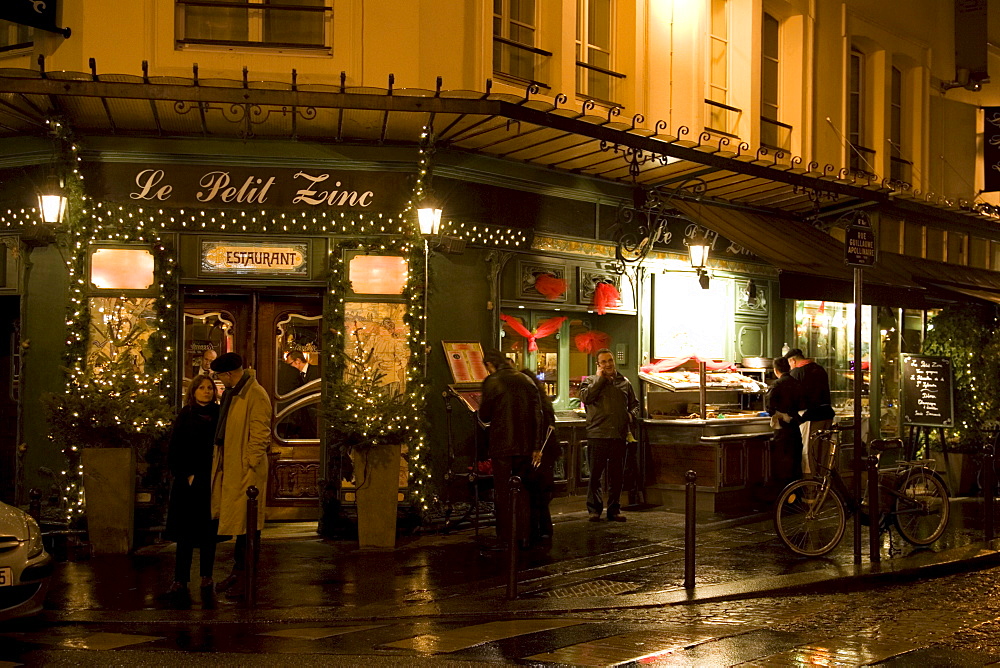 People shelter from rain at night on street corner outside traditional Le Petit Zinc Restaurant, Left Bank, Paris, France