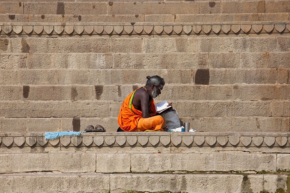 Hindu Sadhu holy man with traditional robe reads on steps of the Ghats in holy city of Varanasi, Benares, Northern India