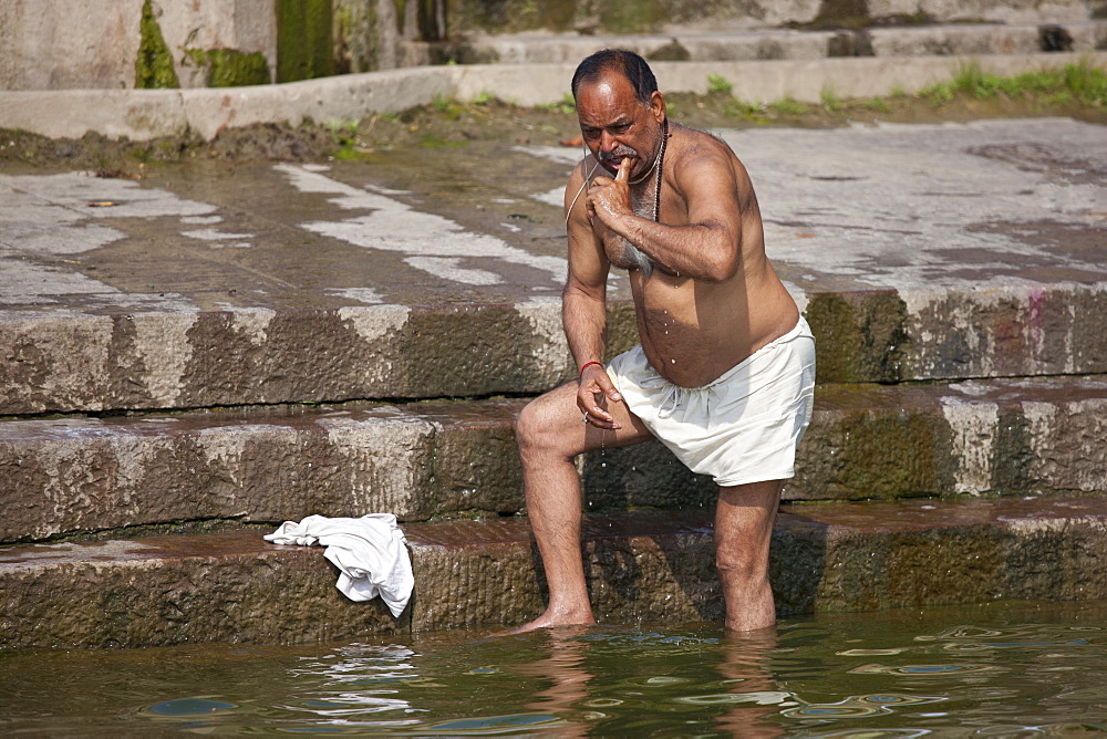 Indian Hindu man bathing and cleaning his teeth in the River Ganges by Kshameshwar Ghat in holy city of Varanasi, India