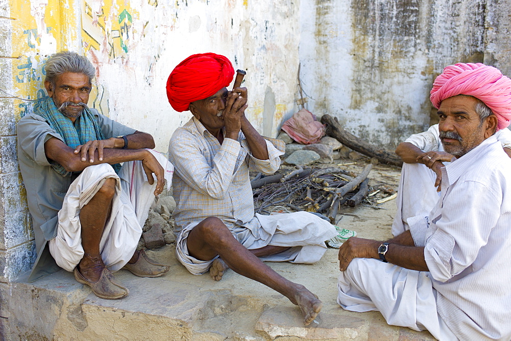 Indian man in traditional clothing smokes clay pipe while sitting with friends in Narlai village in Rajasthan, Northern India