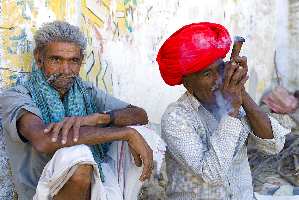 Indian man in traditional clothing smokes clay pipe while sitting with a friend in Narlai village in Rajasthan, Northern India