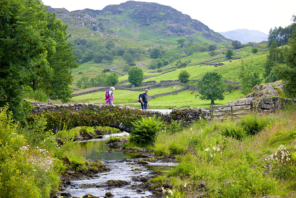 Walkers cross Packhorse Bridge across mountain stream at Watendlath in the Lake District National Park, Cumbria, UK