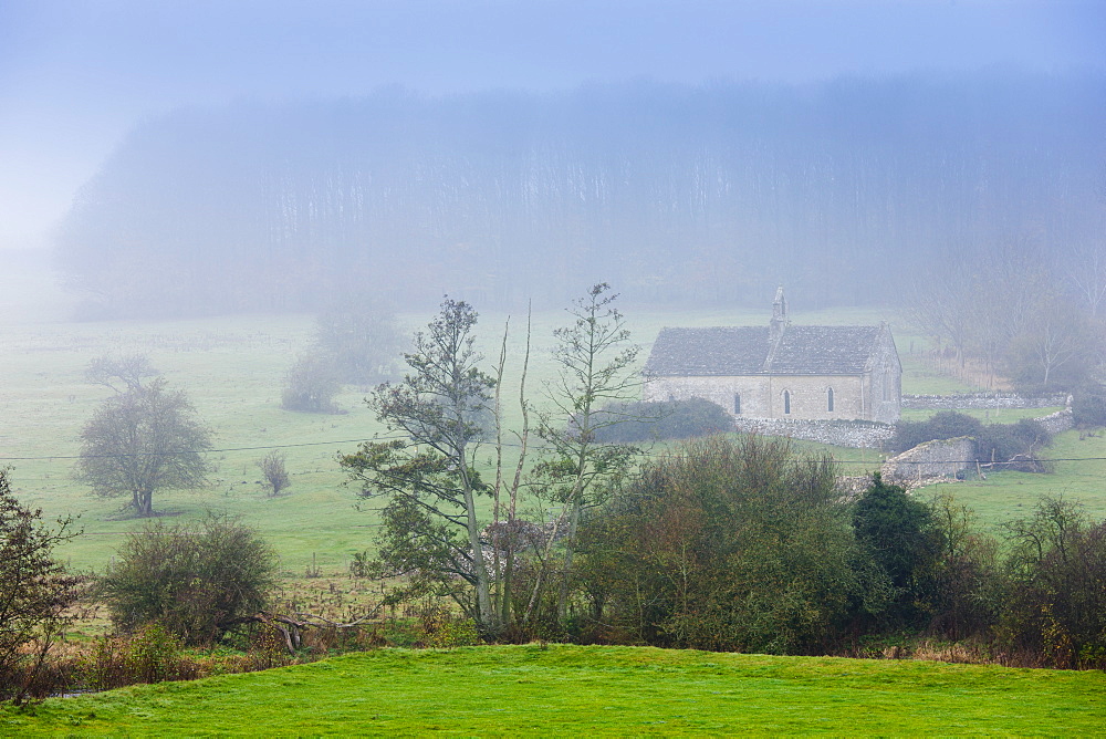 Misty scene and St Oswald's Church, a remote chapel in a field, at Widford near Burford in The Cotswolds, Oxfordshire, UK