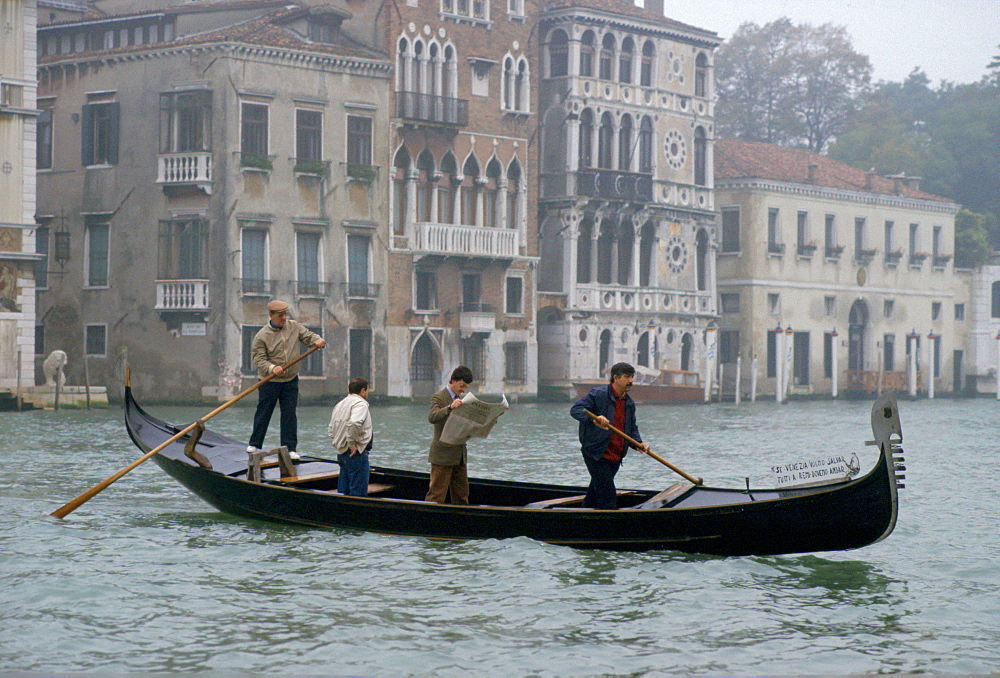 On a grey day in Venice a man commuting by gondola on the canal stands up to read his newspaper.