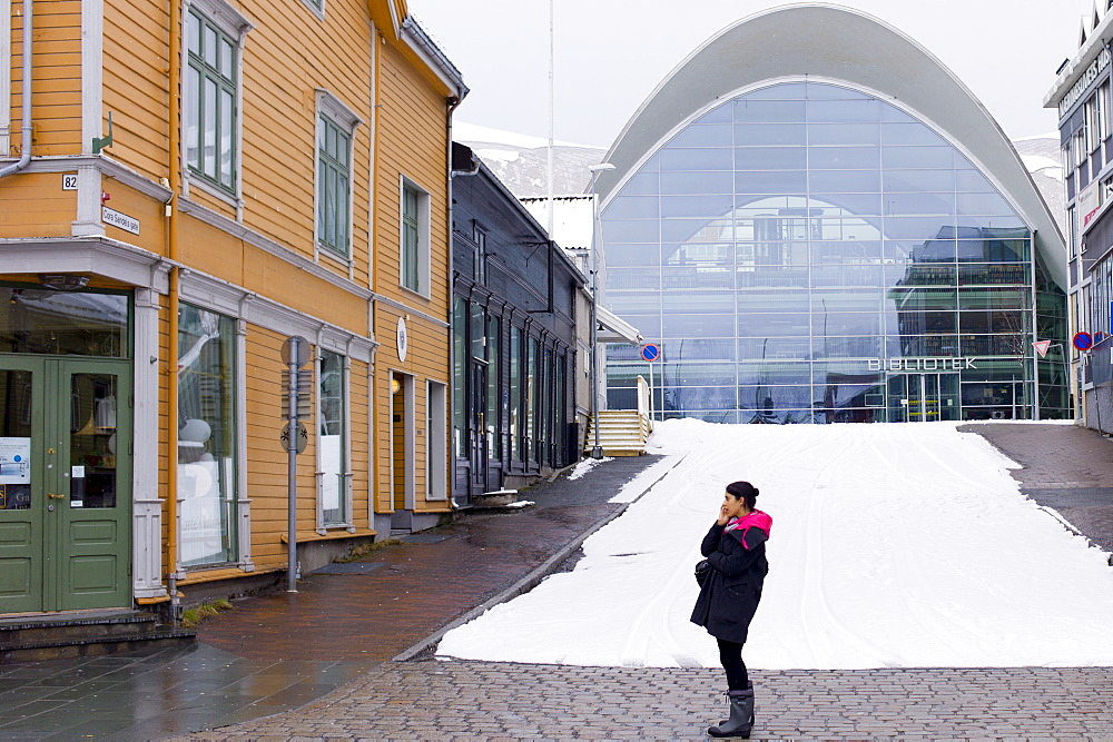 Local woman using mobile phone in street scene by modern Biblioteket library in Tromsoya, Tromso, in Northern Norway