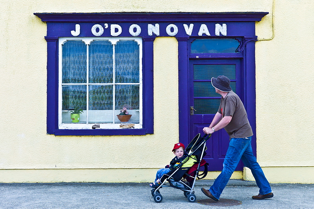 Man wheels child in stroller past J'ODonovan shop front at Courtmacsherry, County Cork, Ireland