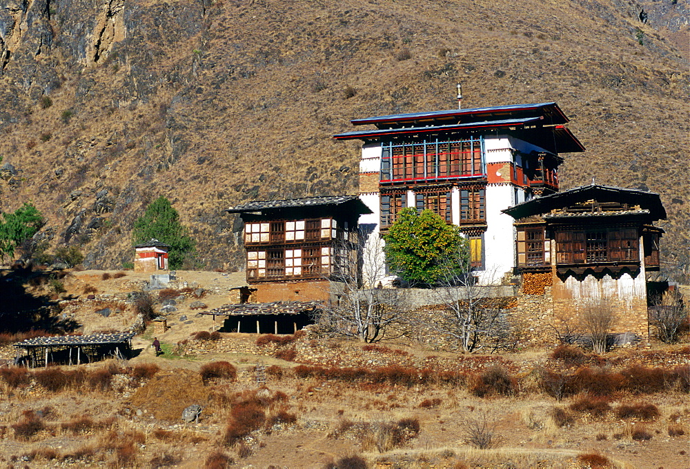 A typical Bhutanese home built in a traditional chalet style to suit the alpine surroundings.  The central building has a raised roof and wooden-shuttered windows. The surrounding land has been terraced with stone walls.