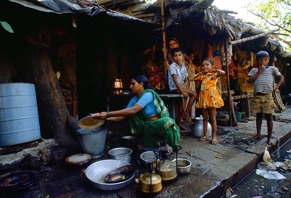 Watched by a group of young children a woman washes dishes in a bucket of water at a humble home on the sidewalk in Bombay, India.  A little girl has a gleeful expression despite the poverty of the situation.