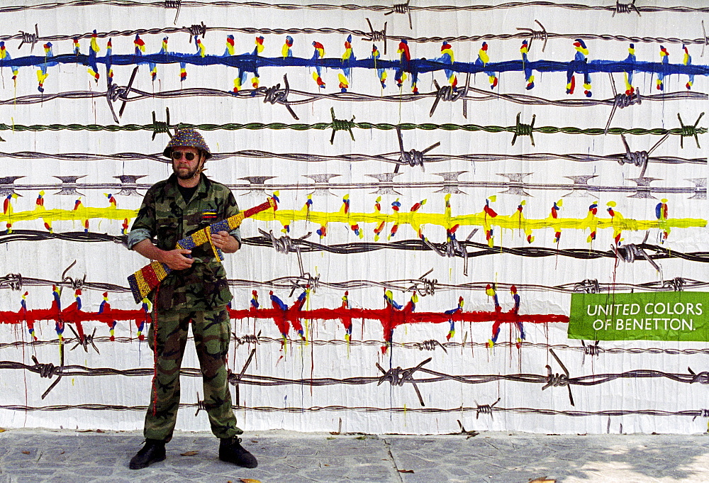 Man dressed as soldier with coloured weapon in front of United Colors of Benetton poster in 1980s in Italy