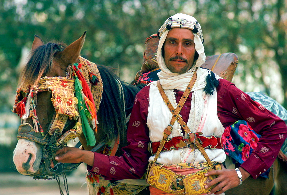 A horseman in traditional dress holding his horse which has eye defenders against the sun and dust.