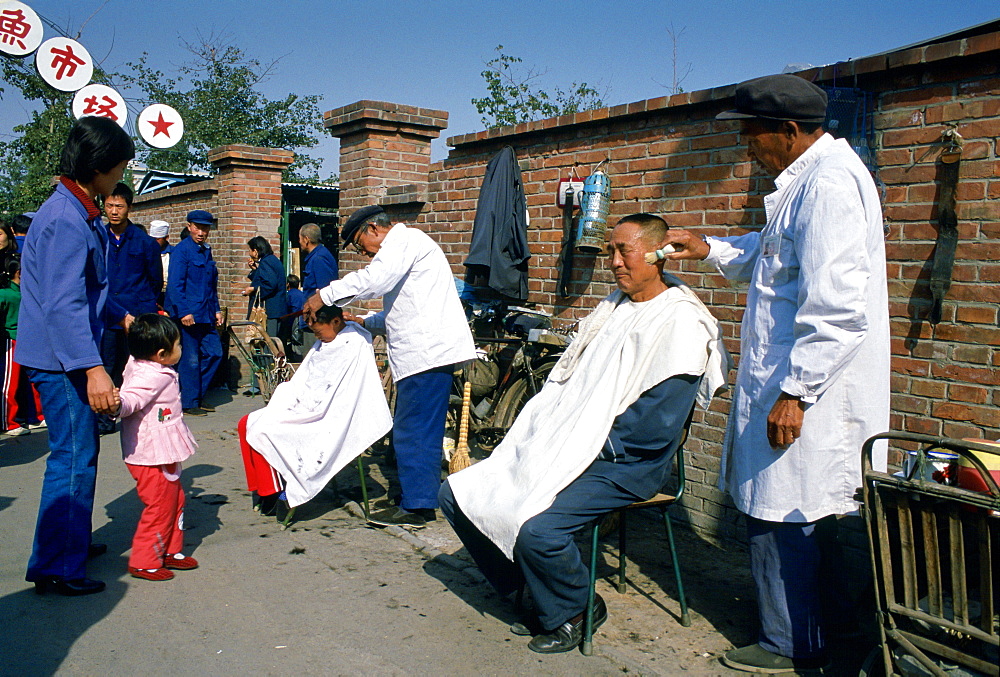 Men at work at an outdoor Barbers' Shop in Old Peking (Beijing), China.  One is using a shaving brush with foam and the other is cutting a young boy's hair watched by a mother and child.