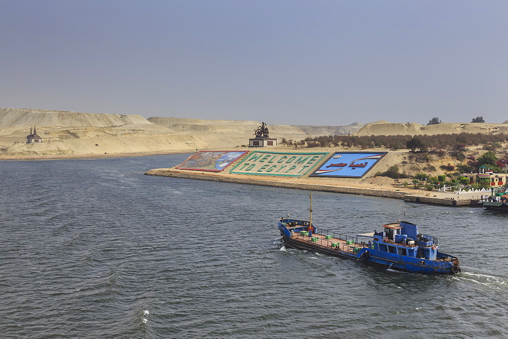 Small ship travels on the Suez Canal, past memorials and a large Welcome to Egypt sign, Ismalia, Egypt, North Africa