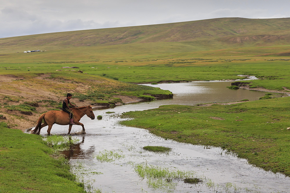 Nomadic boy rides his horse through river with gers int he distance at summer nomad camp, Khujirt, Uvurkhangai (Ovorkhangai), Central Mongolia, Central Asia, Asia